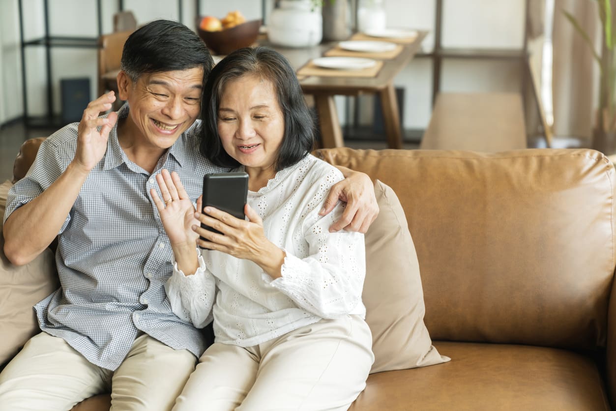 Happy senior old asian lover couple holding smartphone looking at cellphone screen laughing casual relaxing sit on sofa together, smiling elder mature grandparents family embracing lifestyle