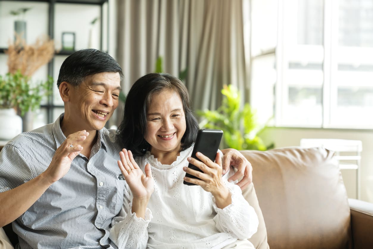 Happy senior old asian lover couple holding smartphone looking at cellphone screen laughing casual relaxing sit on sofa together, smiling elder mature grandparents family embracing lifestyle